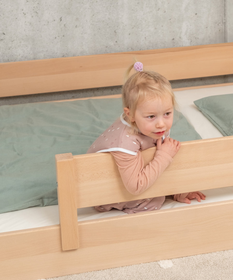 Sicher & geborgen*Mit dem abnehmbaren Rausfallschutz bietet das Bett schon kleineren Kindern Sicherheit und sorgt für extra Geborgenheit.|Mädchen sitzt am Rausfallschutz von Bodenbett Buche B3 Natur.