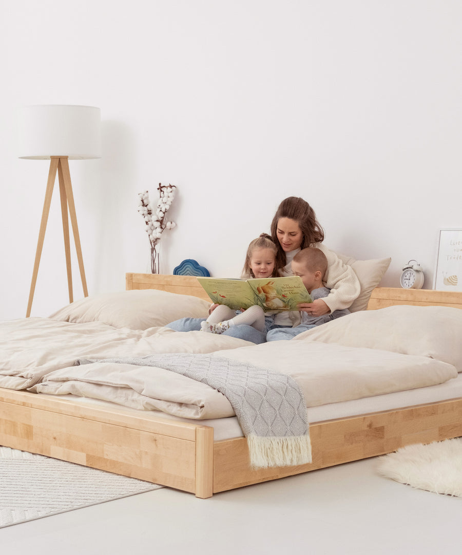 Das perfekte Familienbett*Flexibel dank modularer Bauweise und passend für jedes Zuhause: Unser Bett ist in zwei Höhen und zwei Größen erhältlich.|Mutter mit Kindern schaut Bilderbuch an auf Familienbett Birke bodentief.
