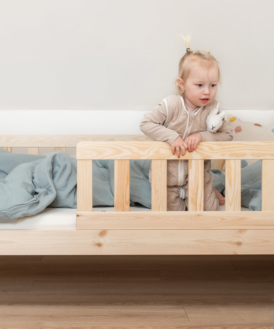 Flexibler Rausfallschutz*Der Rausfallschutz bietet deinem Kind Sicherheit. Er lässt sich beidseitig montieren und bei Bedarf auch ganz abnehmen.|Kind sitzt in Kinderbett Kiefer Natur und schaut über den praktischen Rausfallschutz.