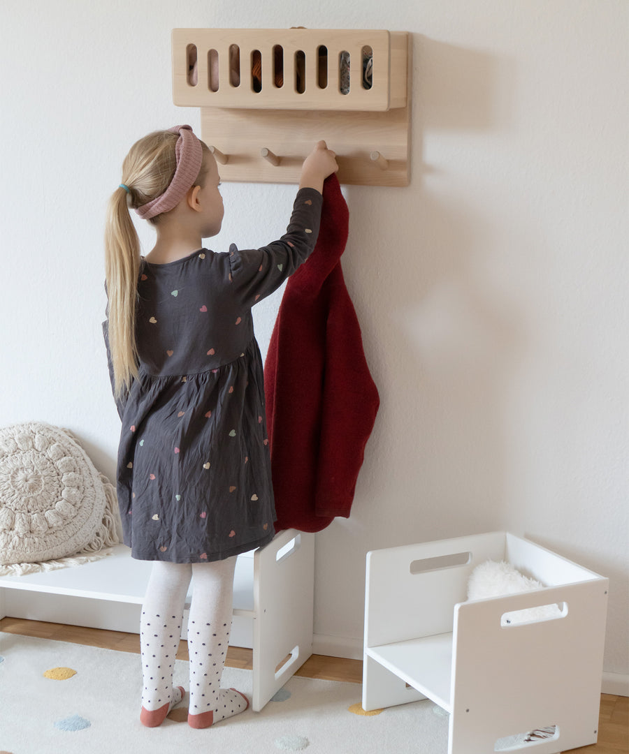 Inspiriert von Montessori*Durch das praktische Ablagefach, das sich auf Kinderhöhe befindet, kann dein Kind selbstständig Ordnung halten.|Mädchen hängt Jacke an Kindergarderobe.