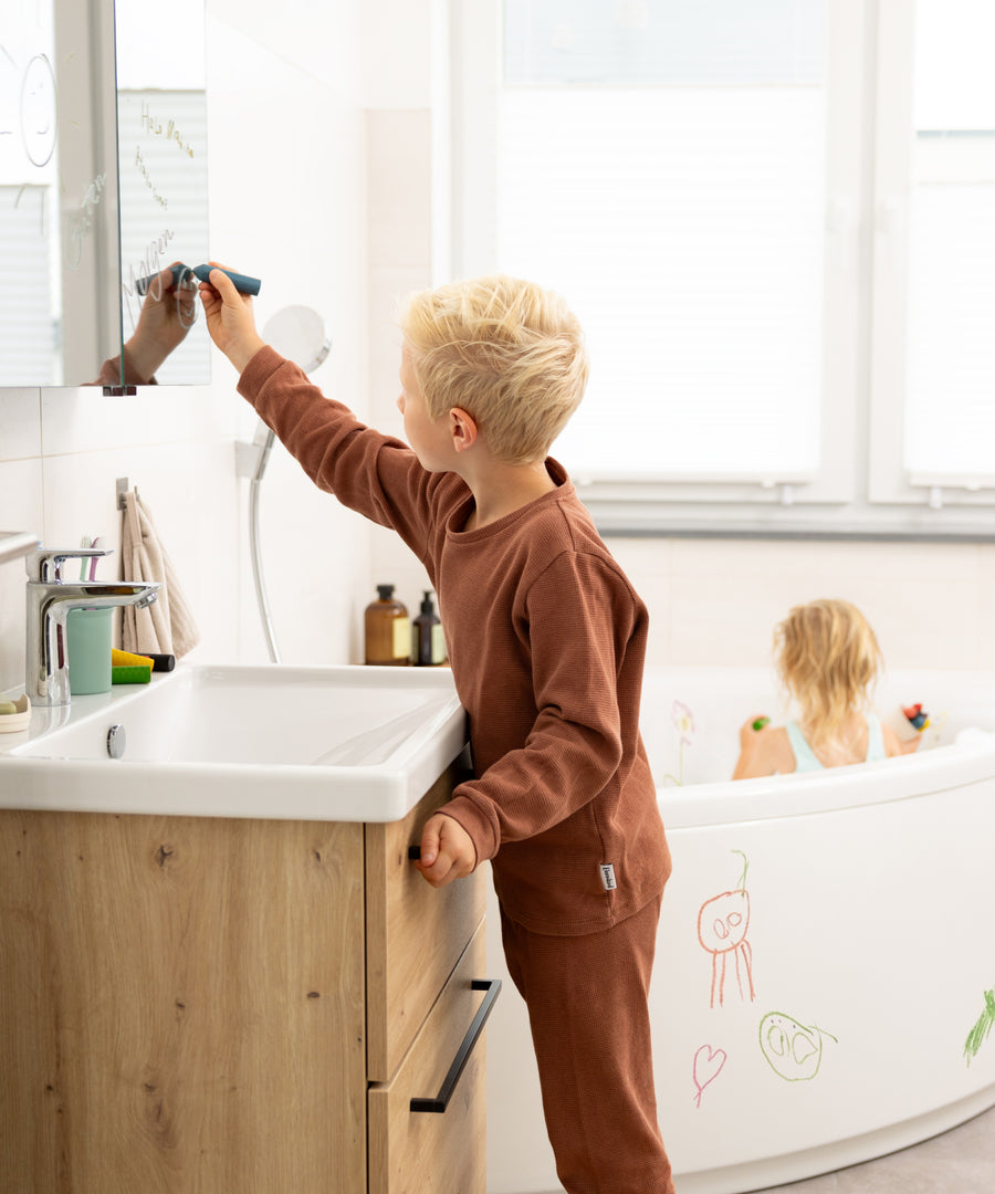 Kind malt mit dem Badewannenstift auf den Spiegel im Badezimmer.