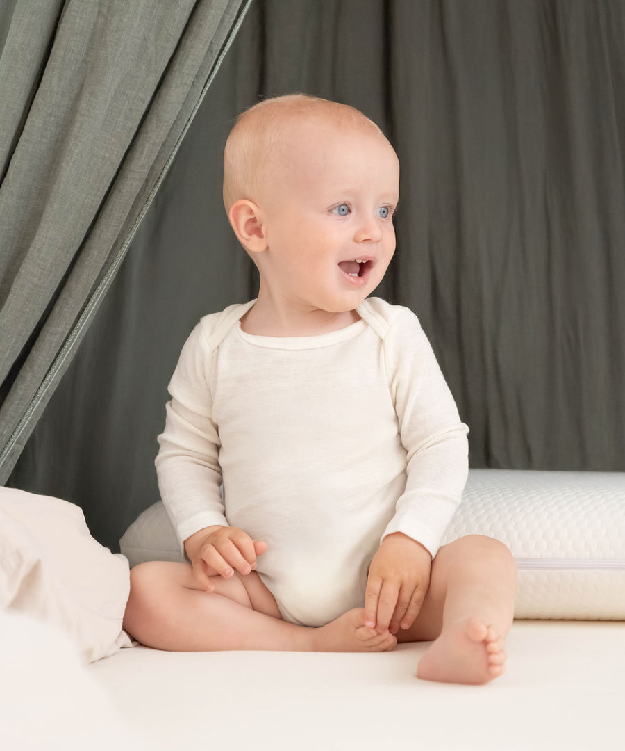 Klimaregulierend*Merinowolle reguliert Feuchtigkeit, hält die Haut warm und trocken und kann so Hautausschlag bei Babys vorbeugen.|Baby sitzt lachend auf Bett.