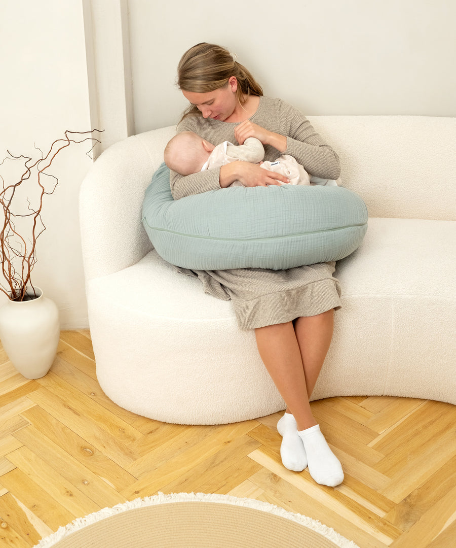 Frau stillt Babymit Hilfe des Stillkissens auf einem Sofa.