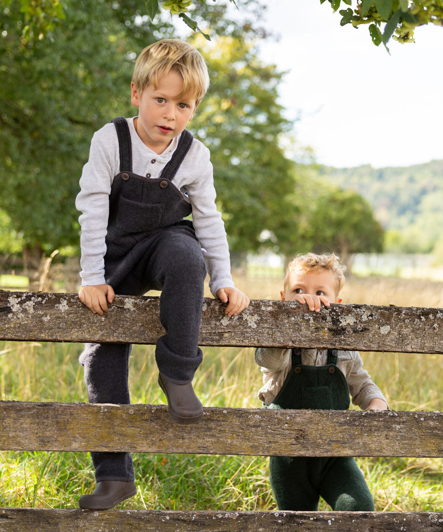 Fair produziert in Europa*Unsere Walk-Latzhose wird mit natürlicher Bio-Schurwolle aus mulesingfreier, kontrolliert biologischer Tierhaltung gefertigt.|Kinder tragen die Walk-Latzhose in Anthrazit und Waldgrün.