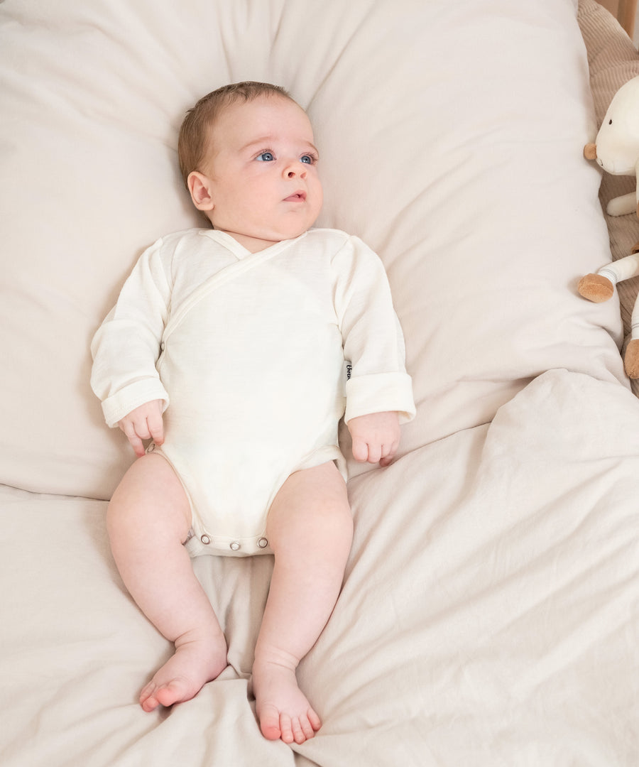 Praktischer Wickelbody*Durch die seitliche Knöpfung und den Wickelschnitt sehr leicht anzuziehen. Die nickelfreien Druckknöpfe sind für Babyhaut geeignet.|Baby liegt auf Bett.